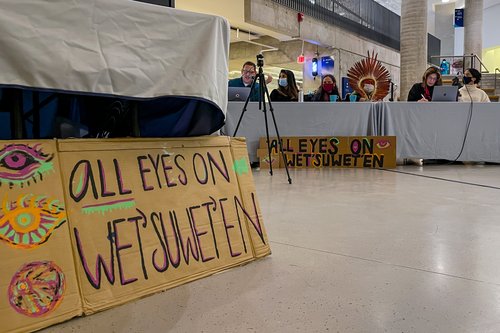 A table with a panel of speakers at the town hall in the background, behind a hand-painted cardboard sign that says "All eyes on Wet'suwet'en"